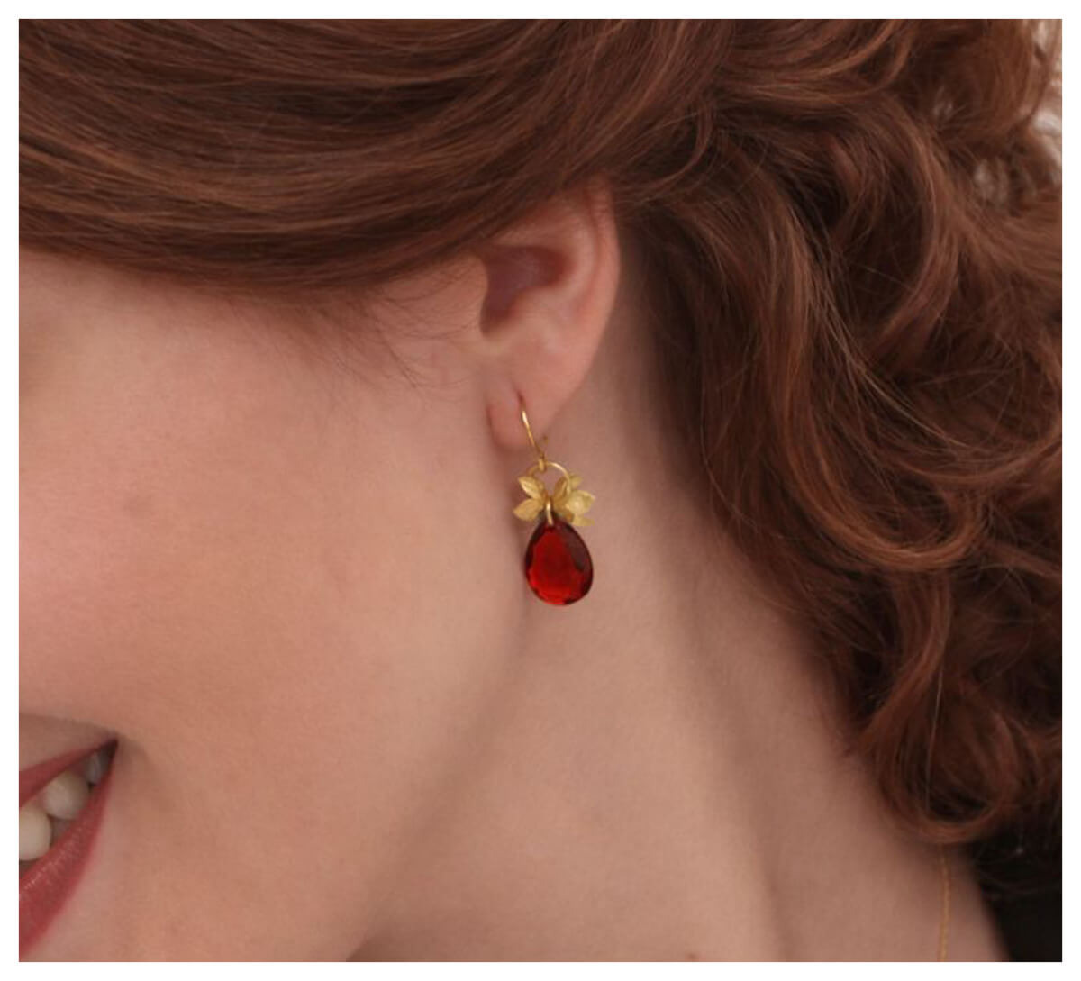 Preview: Nahaufnahme eines Ohrs mit dem Ohrring „Frieda“ mit 750er Goldauflage, gestaltet als kleine Blüten mit einem facettiertem, dunkelroten Tropfenanhänger.